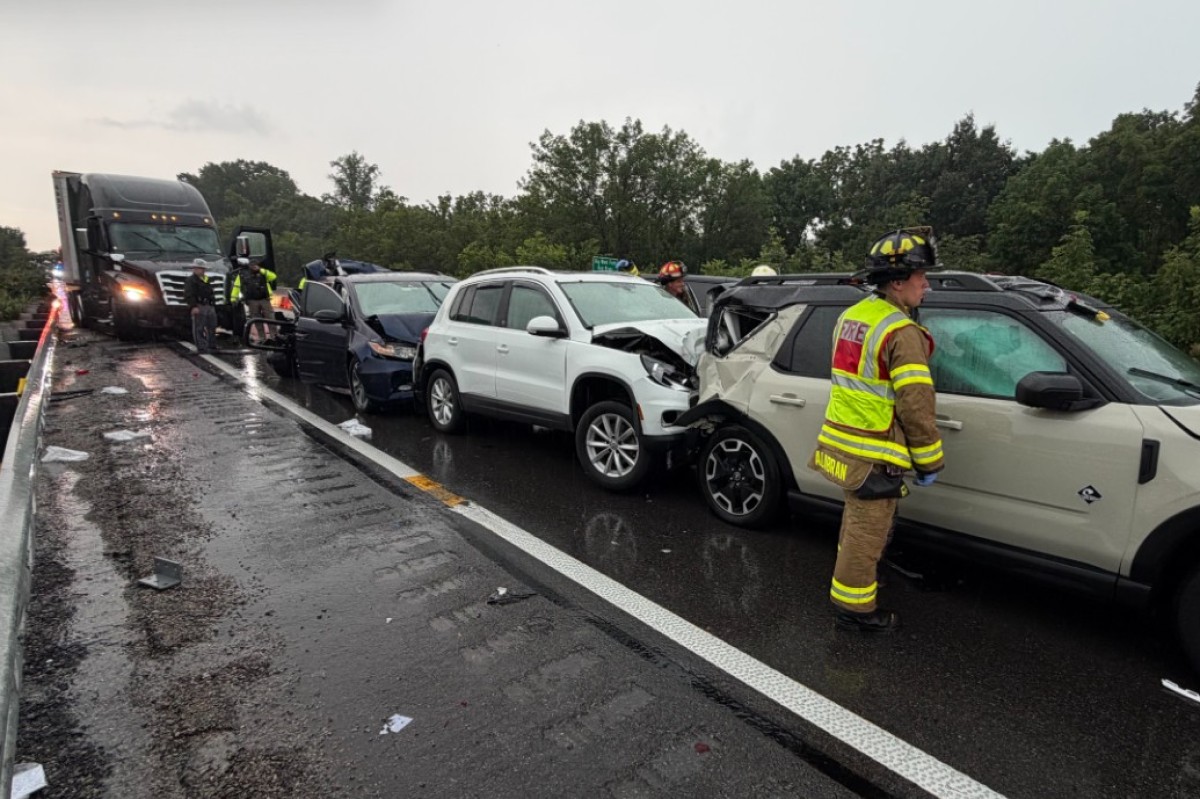 Multi‑vehicle crash on a rainy highway: a white SUV and a beige SUV show front‑end damage, backed by a dark blue car, with a tow truck arriving and firefighters in reflective gear on the scene.