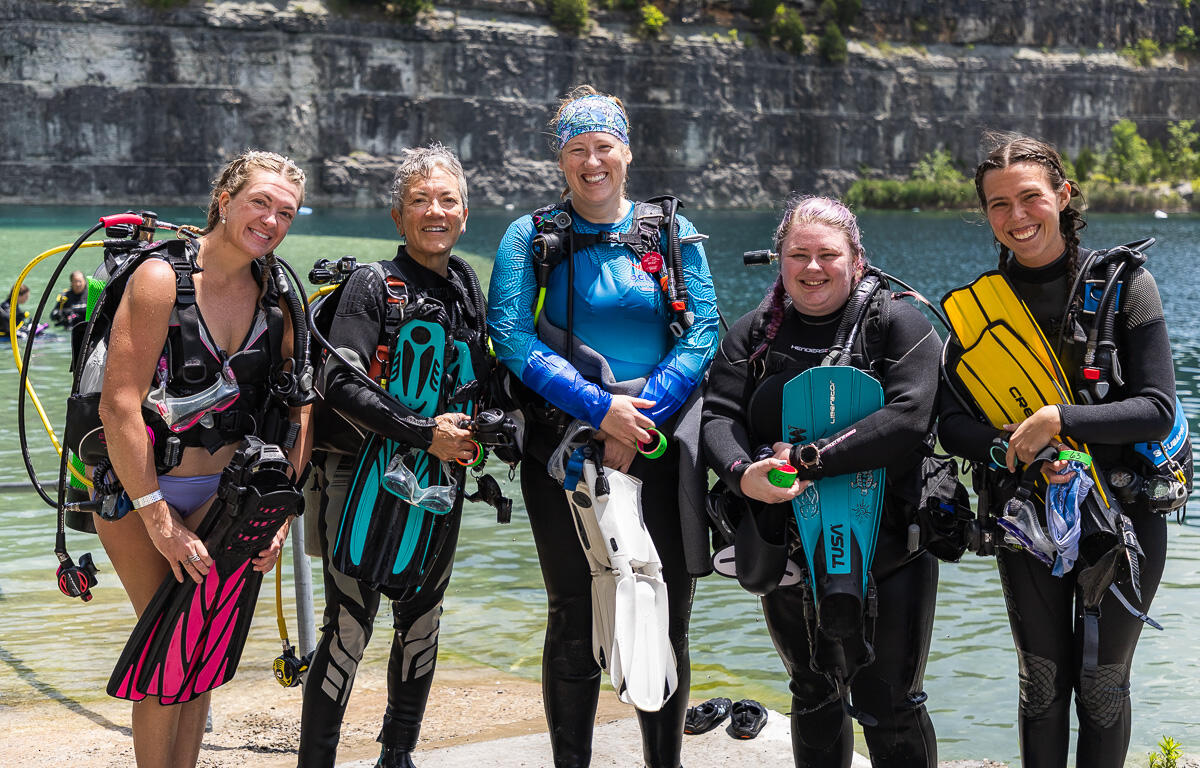 Five female scuba divers stand on a lakeshore in full gear, smiling at the camera.