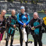 Five female scuba divers stand on a lakeshore in full gear, smiling at the camera.