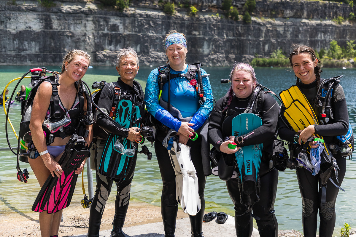 Five female scuba divers stand on a lakeshore in full gear, smiling at the camera.