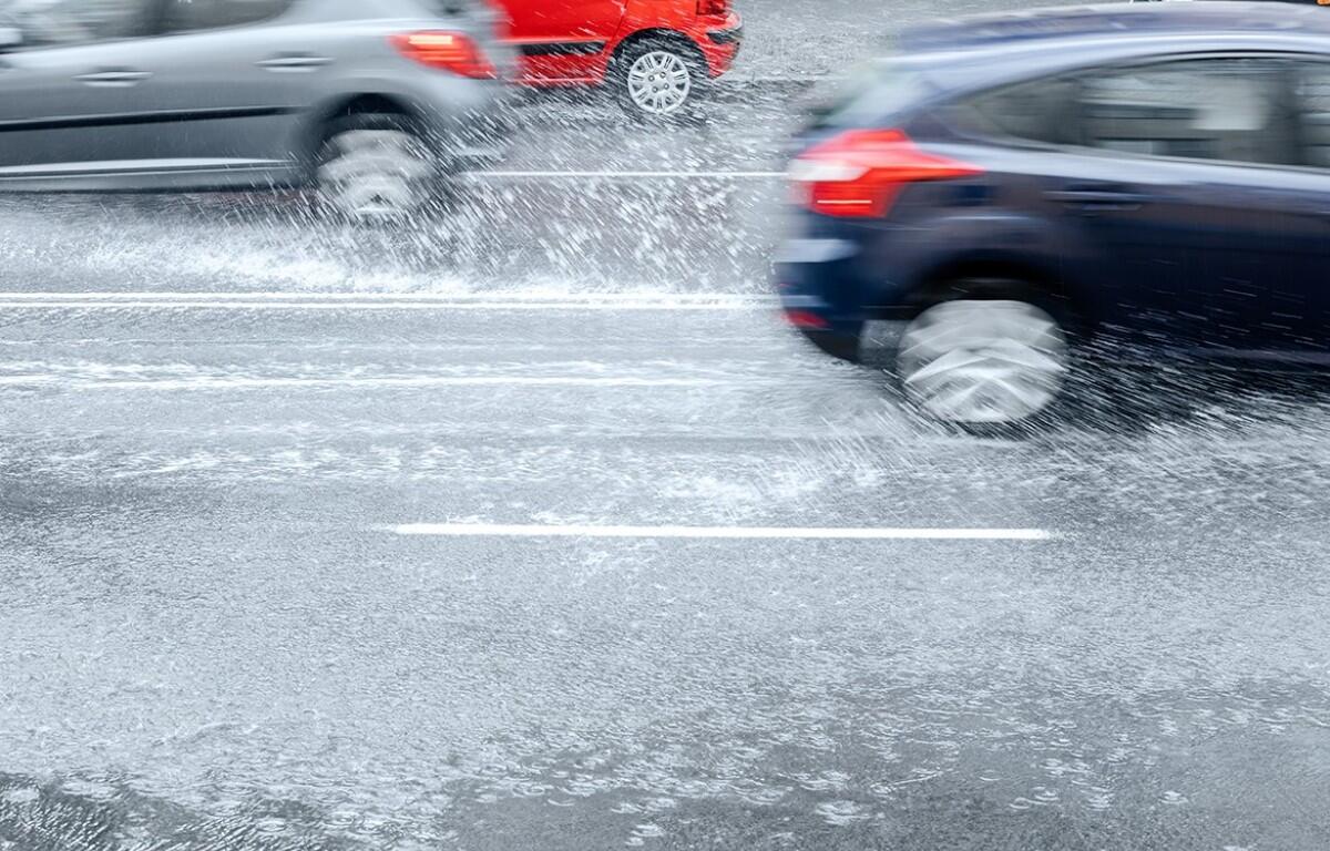 Cars drive through a flooded road, spraying water as they pass.