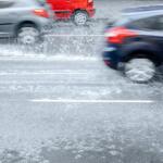 Cars drive through a flooded road, spraying water as they pass.