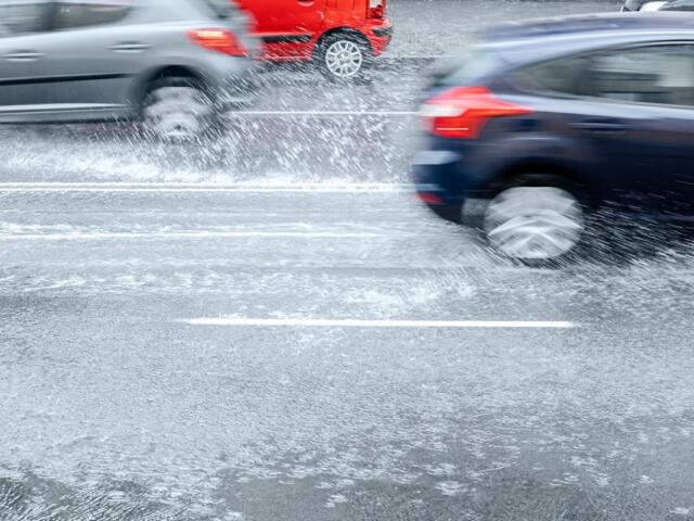 Cars drive through a flooded road, spraying water as they pass.