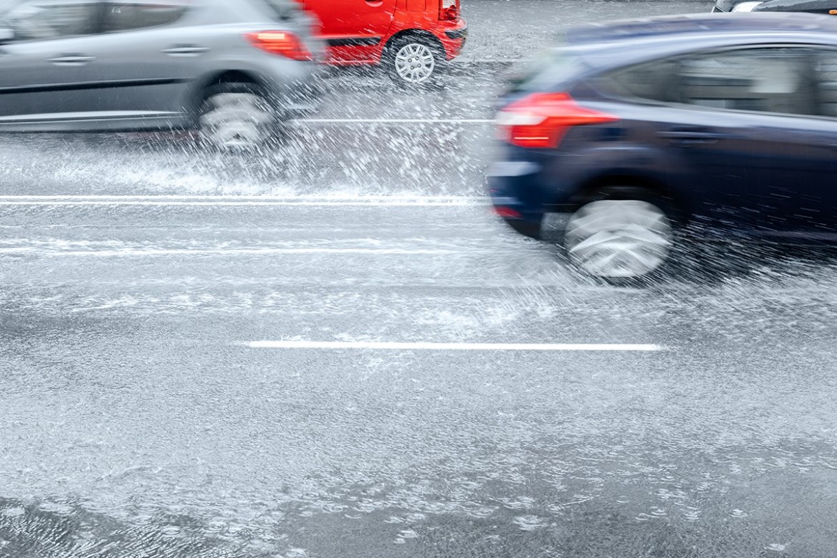 Cars drive through a flooded road, spraying water as they pass.