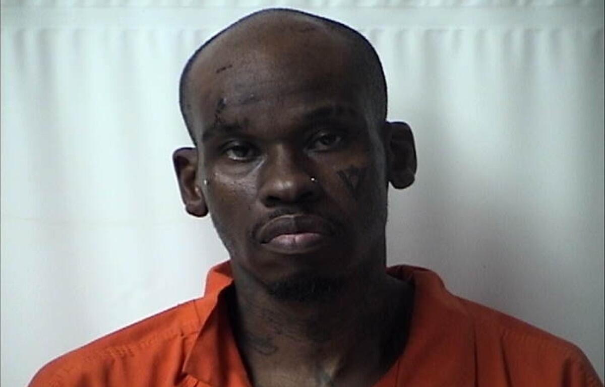 Mugshot of a dark-skinned man in an orange jail jumpsuit against a light background, with visible facial tattoos and a solemn expression.