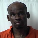 Mugshot of a dark-skinned man in an orange jail jumpsuit against a light background, with visible facial tattoos and a solemn expression.