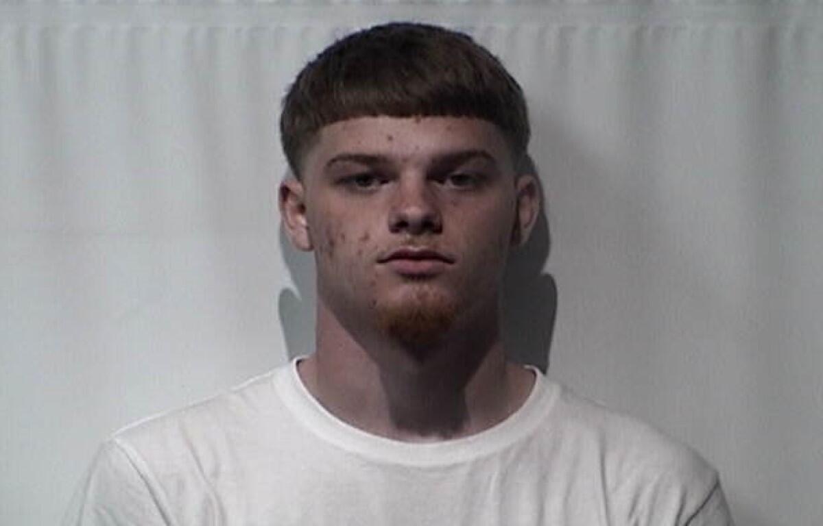 Mugshot-style photo of a young man with short dark hair and a slight goatee, wearing a white t-shirt against a plain backdrop.