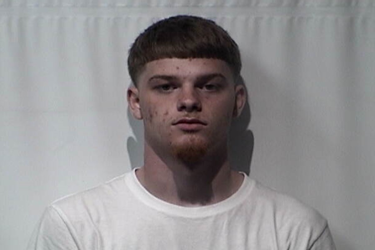 Mugshot-style photo of a young man with short dark hair and a slight goatee, wearing a white t-shirt against a plain backdrop.
