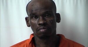 Mugshot of a dark-skinned man in an orange jail jumpsuit against a light background, with visible facial tattoos and a solemn expression.