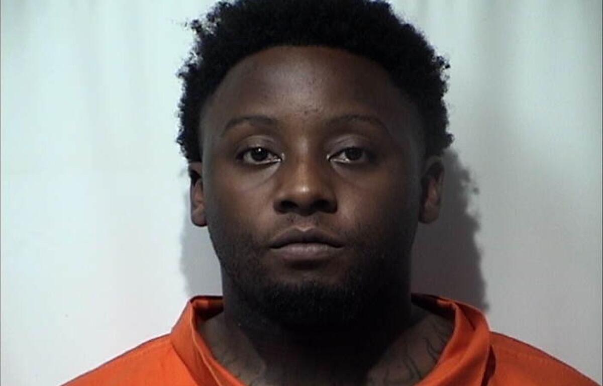 Mugshot of a Black man with short curly hair in an orange inmate jumpsuit against a light background, neutral expression.