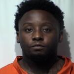 Mugshot of a Black man with short curly hair in an orange inmate jumpsuit against a light background, neutral expression.