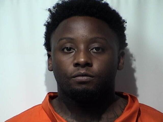 Mugshot of a Black man with short curly hair in an orange inmate jumpsuit against a light background, neutral expression.
