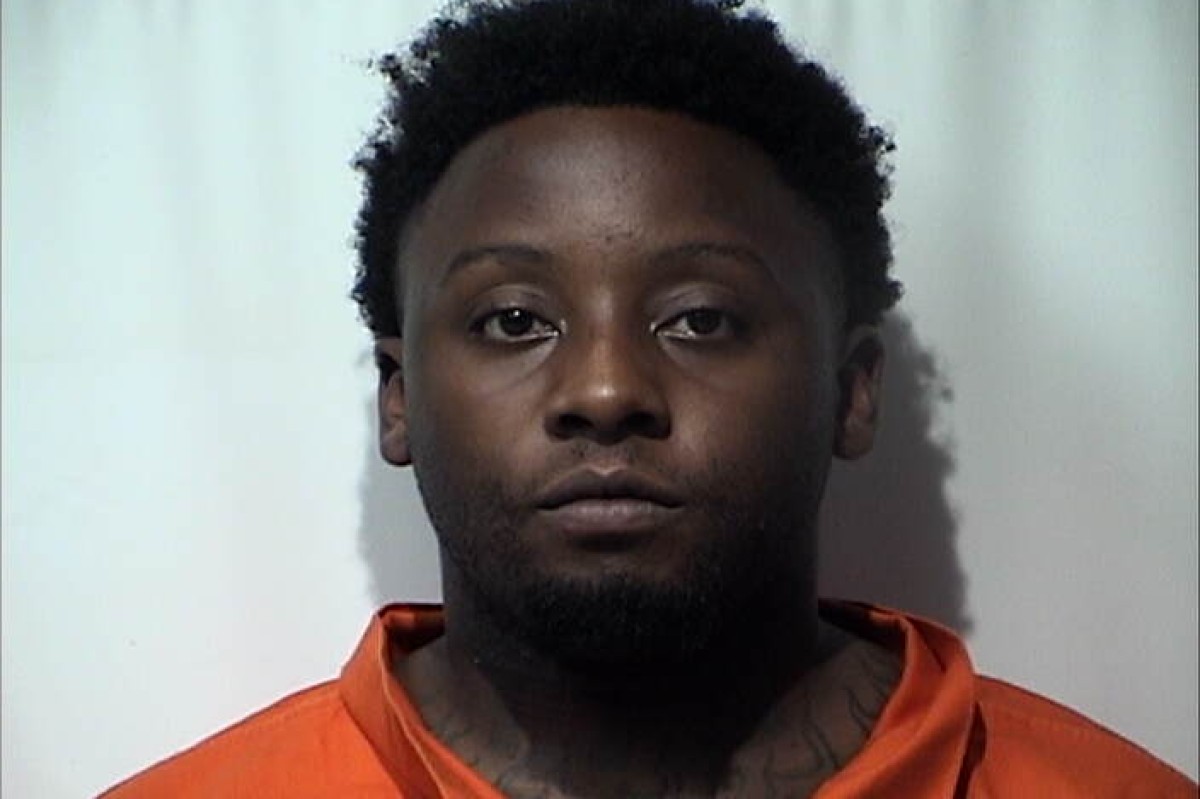 Mugshot of a Black man with short curly hair in an orange inmate jumpsuit against a light background, neutral expression.