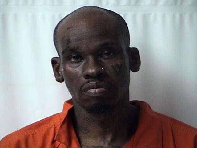 Mugshot of a dark-skinned man in an orange jail jumpsuit against a light background, with visible facial tattoos and a solemn expression.