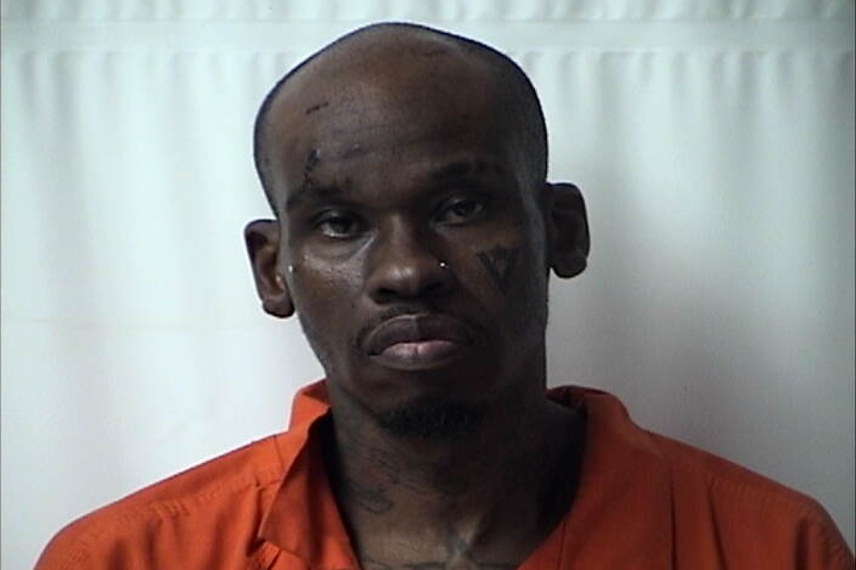 Mugshot of a dark-skinned man in an orange jail jumpsuit against a light background, with visible facial tattoos and a solemn expression.