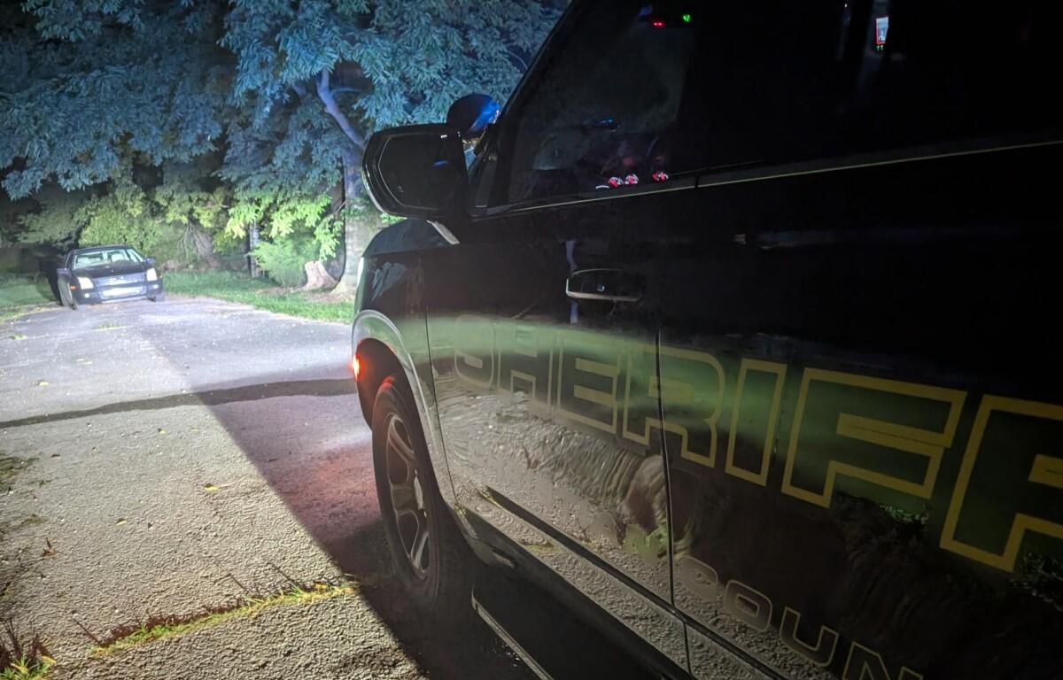 Close-up of a dark sheriff SUV at night, showing yellow lettering on the side and a red taillight.