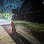 Close-up of a dark sheriff SUV at night, showing yellow lettering on the side and a red taillight.
