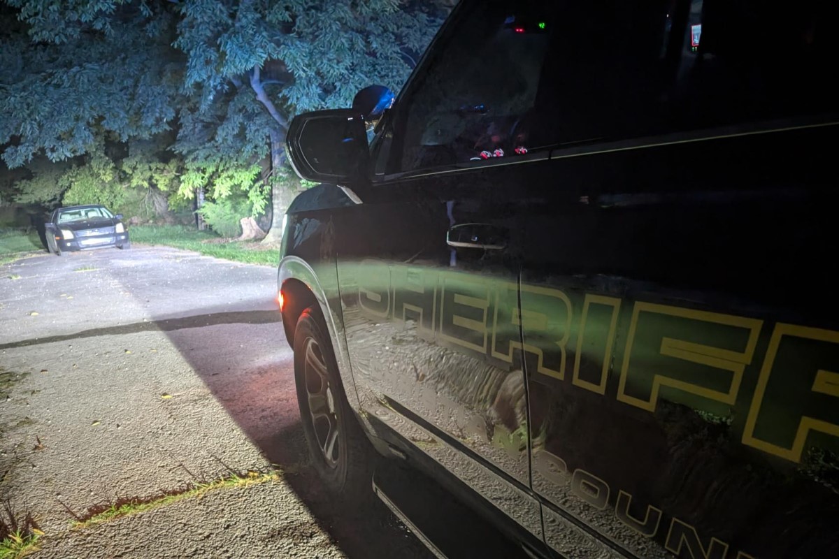 Close-up of a dark sheriff SUV at night, showing yellow lettering on the side and a red taillight.